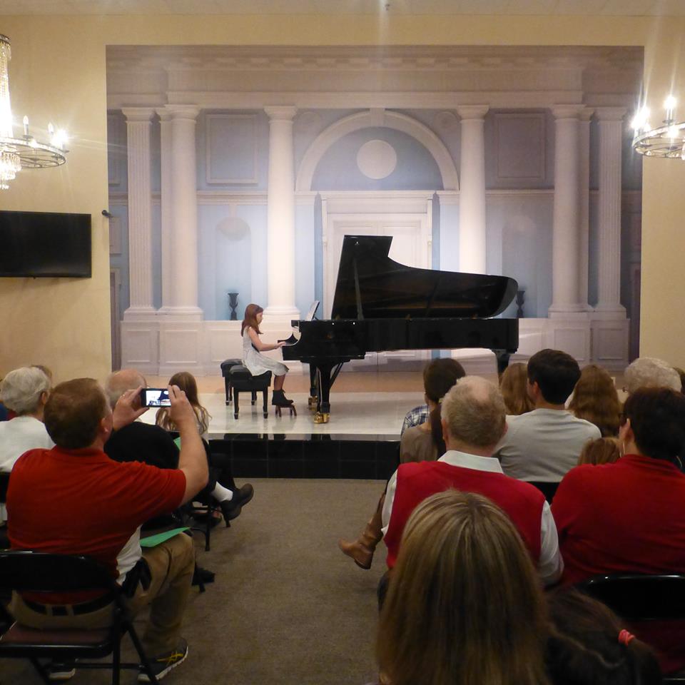 picture shows a recital hall full of people with a student at the piano, viewed from the back of the hall